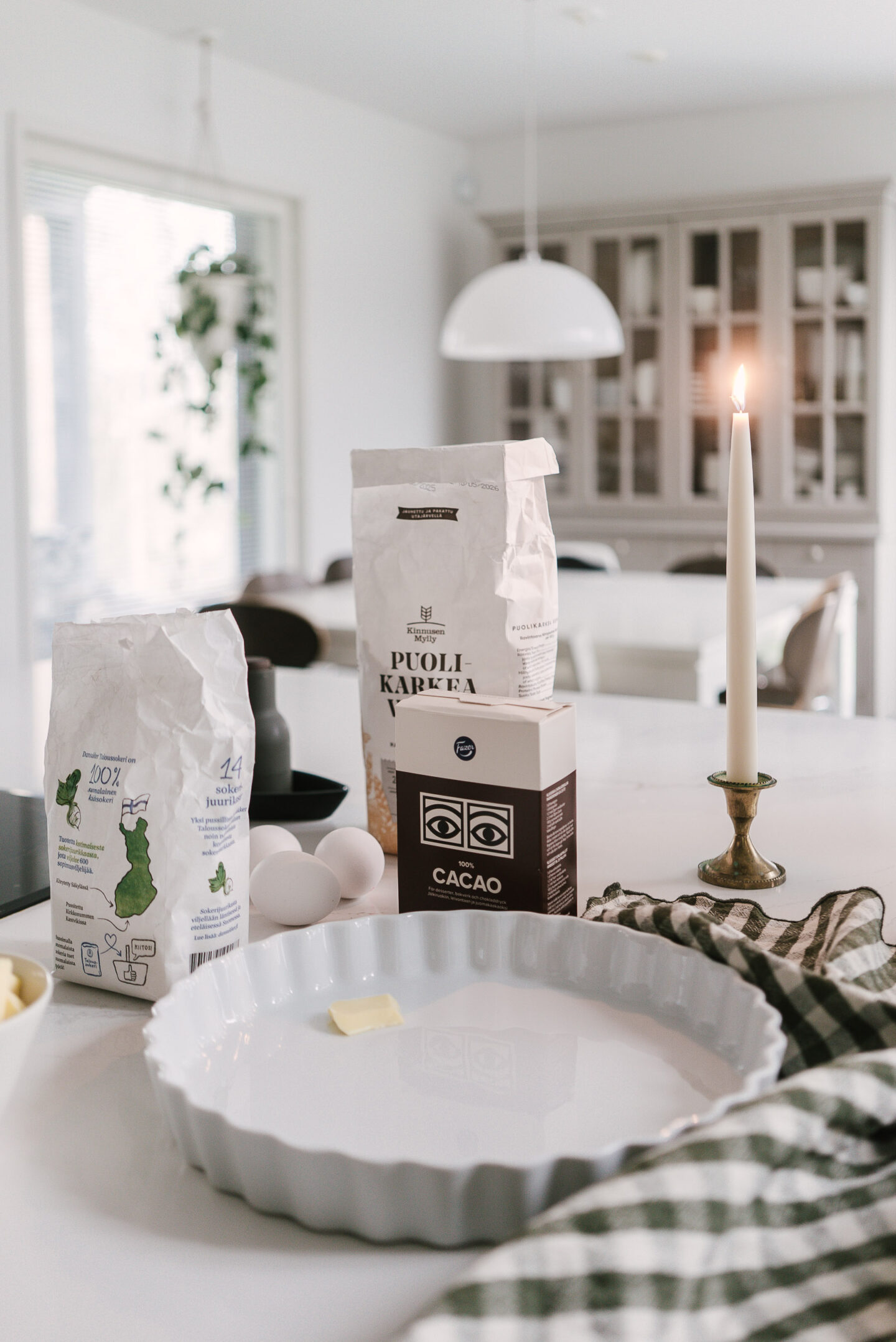 Helpon mutakakun ainekset keittiön saarekkelle nostettuna: jauhopussi, kaakaopaketti, sokeripussi, voita ja kananmunia sekä piirakkavuoka.